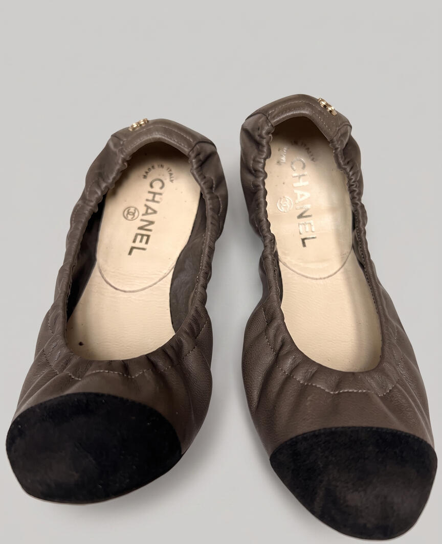 Top-down view of authentic pre-owned Chanel lambskin stretch ballet flats in taupe olive gray leather with black suede cap toes and gold CC logos on a white marble background.