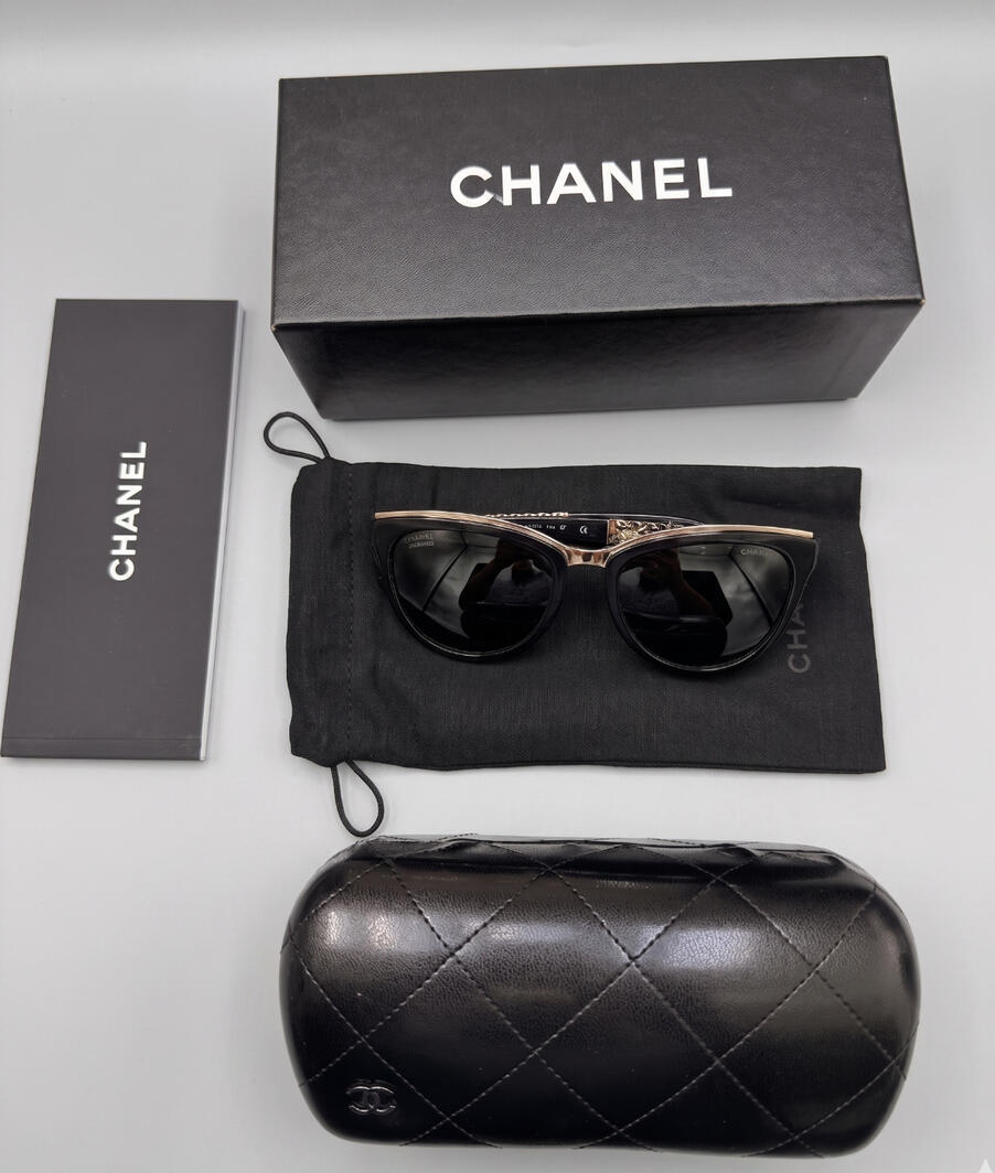 Authentic Chanel 5321 black cat-eye sunglasses featuring gold-tone and black leather chain link arms on a minimalist background.